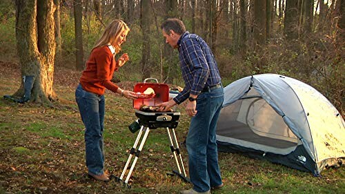 Couple cooking on a portable grill near a tent in the woods.