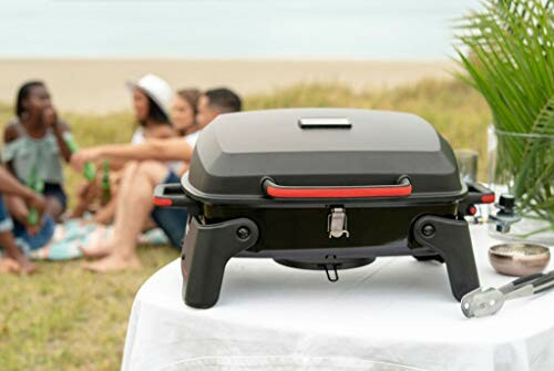 Portable grill on a table with a group of people relaxing on a beach in the background.