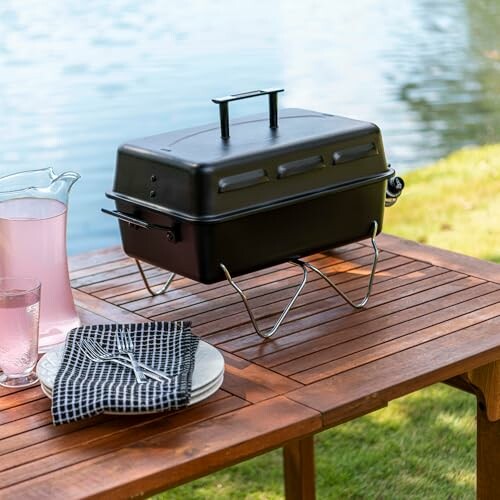 Portable grill on a wooden table by a lake with pink lemonade and plates.